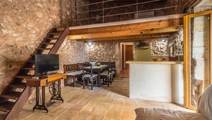 Um interior rústico com uma escadaria de madeira, uma sala de jantar com mesa e cadeiras, uma televisão sobre um móvel de madeira, e uma cozinha com balcão e pia.