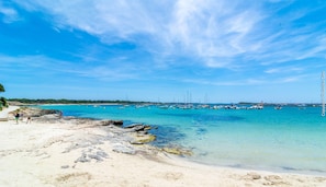 A beach with clear turquoise water, rocky shoreline, and boats in the distance.