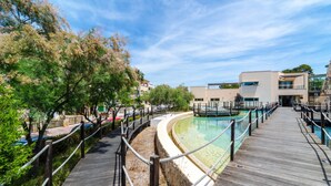 Um calçadão de madeira leva a uma área de piscina com um edifício moderno ao fundo.