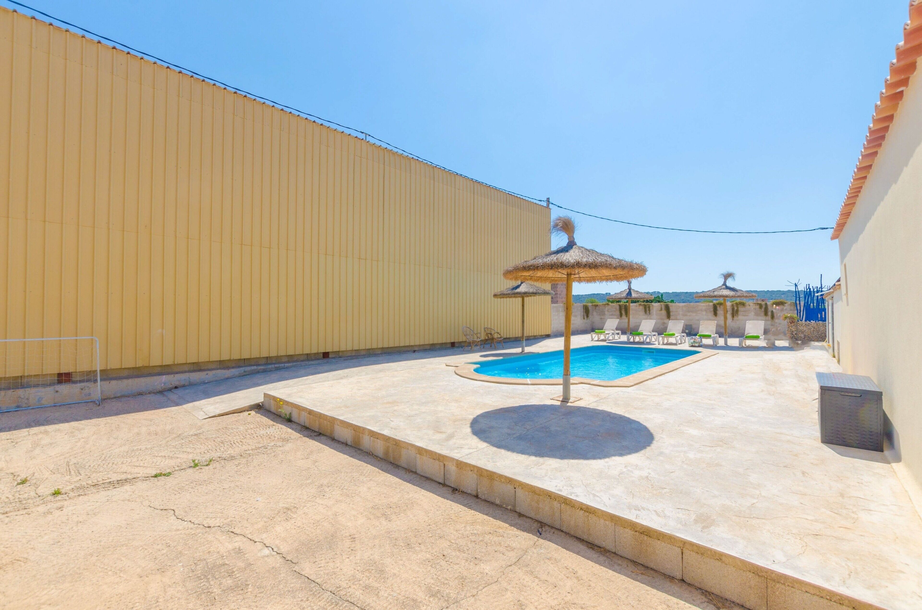 Une piscine entourée de chaises longues et dotée d’un parasol en chaume, le tout bordé d’un mur élevé et surplombé par un ciel dégagé.