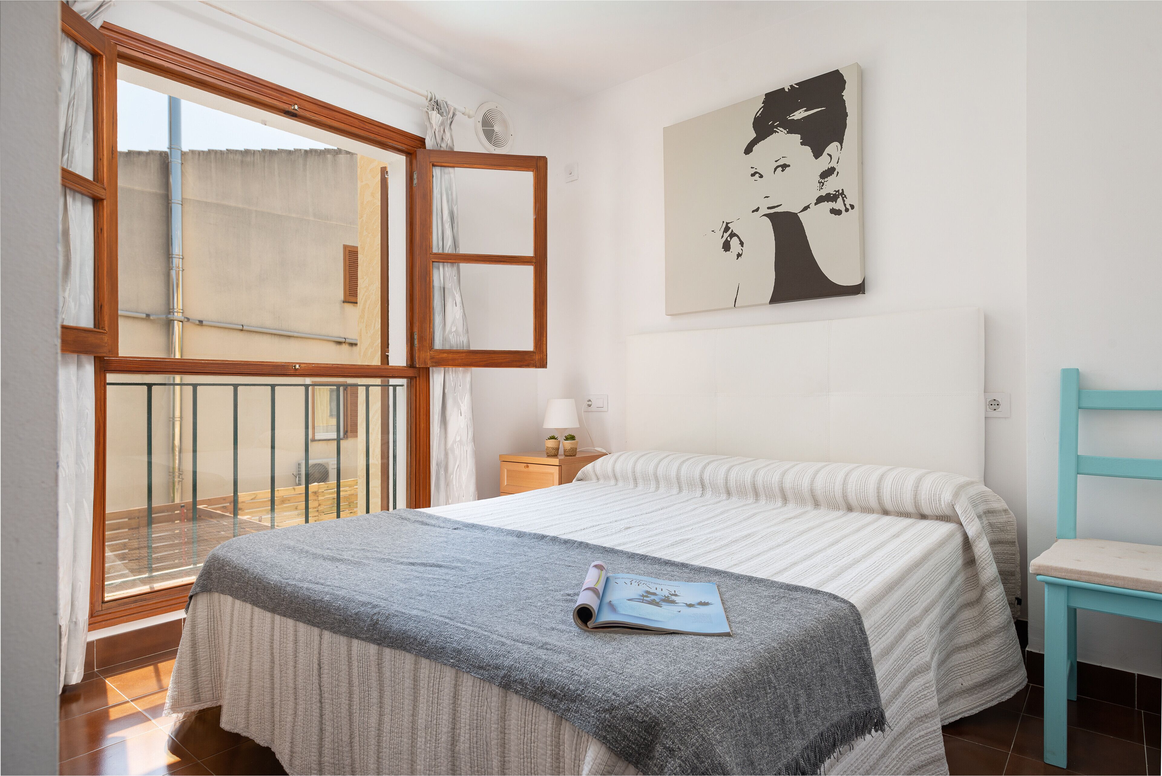 Une chambre à coucher avec un lit, une fenêtre avec un rideau ouvert, une table de chevet avec une lampe et une œuvre d’art accrochée au mur.