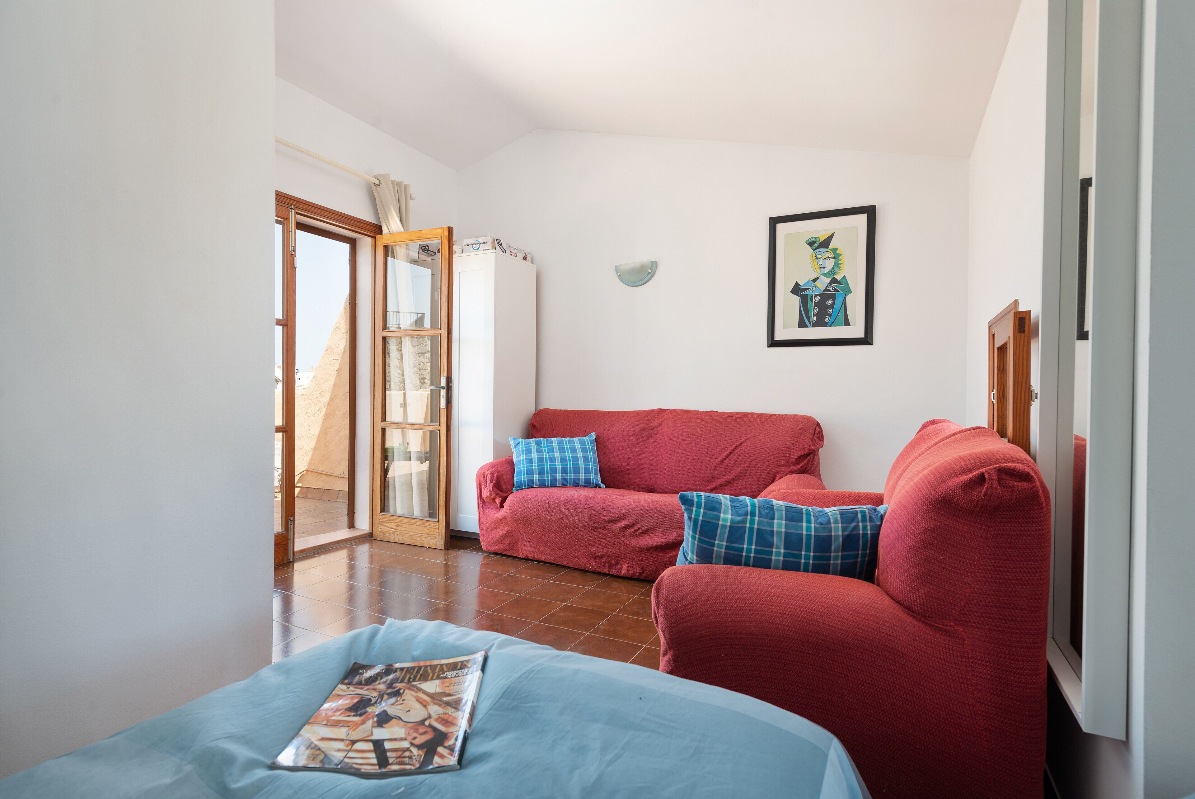 A living room with a red sofa, a blue sofa, and a magazine on a table. There is a door leading to a balcony.
