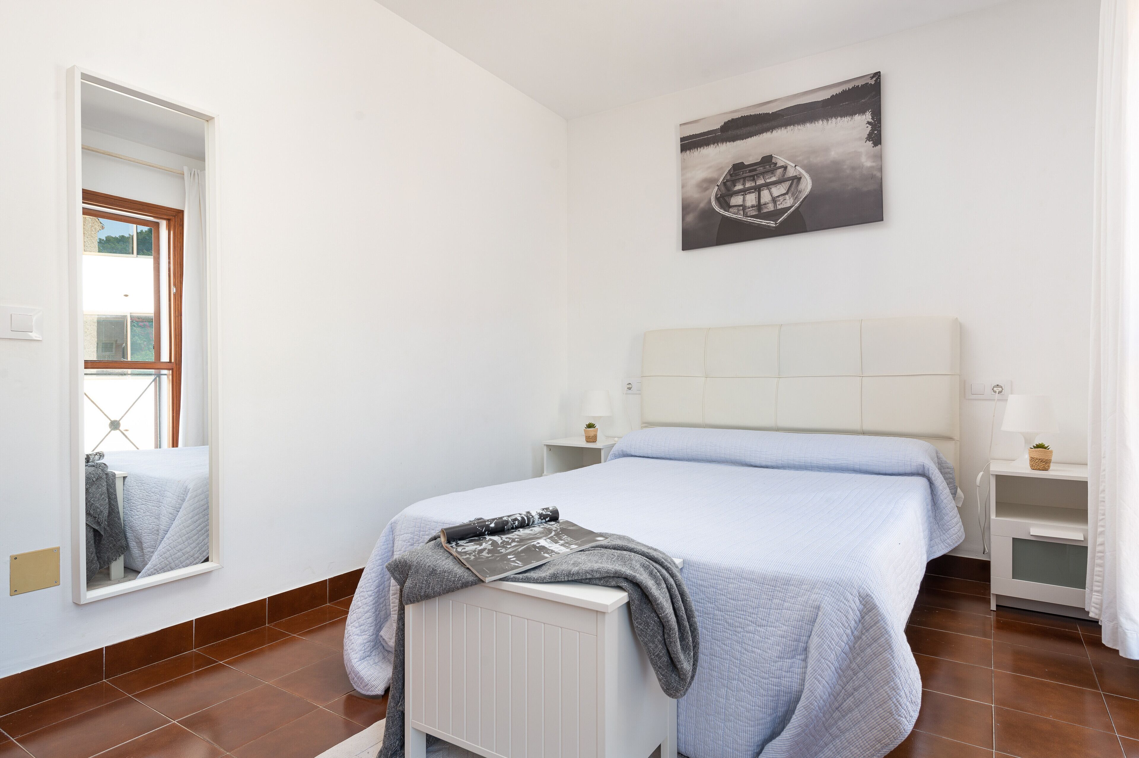 A bedroom with a bed, a bedside table, a nightstand, a window, and a wall-mounted picture.