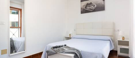A bedroom with a bed, a bedside table, a nightstand, a window, and a wall-mounted picture.