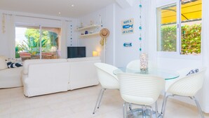 A modern living room with a white sofa, glass dining table, and white chairs. There is a TV on a stand, a hat on the wall, and a sign that says "Have a whale of a time". - BUGAMBILIA - Chalet with terrace in Sa Ràpita. Free WiFi (Sa Ràpita)