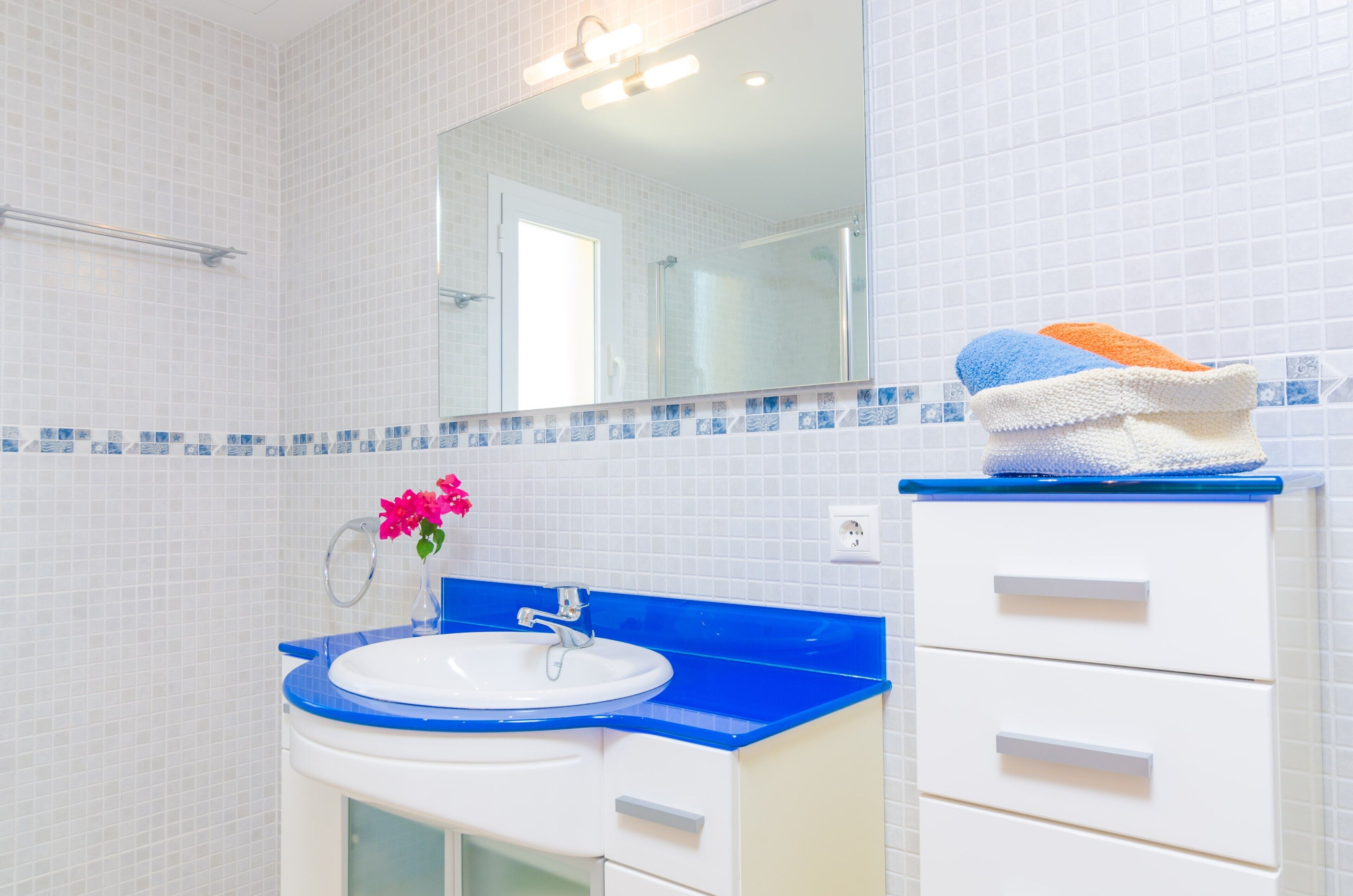Un baño con un lavamanos blanco, encimera azul y un espejo encima. Hay toallas apiladas en un armario, y un florero en la encimera.
