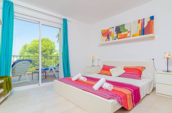 Una camera da letto con un letto, un comodino, una finestra con tende, un balcone con una sedia e un quadro appeso al muro.