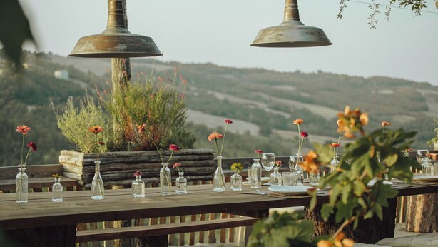 Un área de comedor al aire libre con mesas de madera, jarrones de vidrio transparente con flores y vistas panorámicas a las colinas.