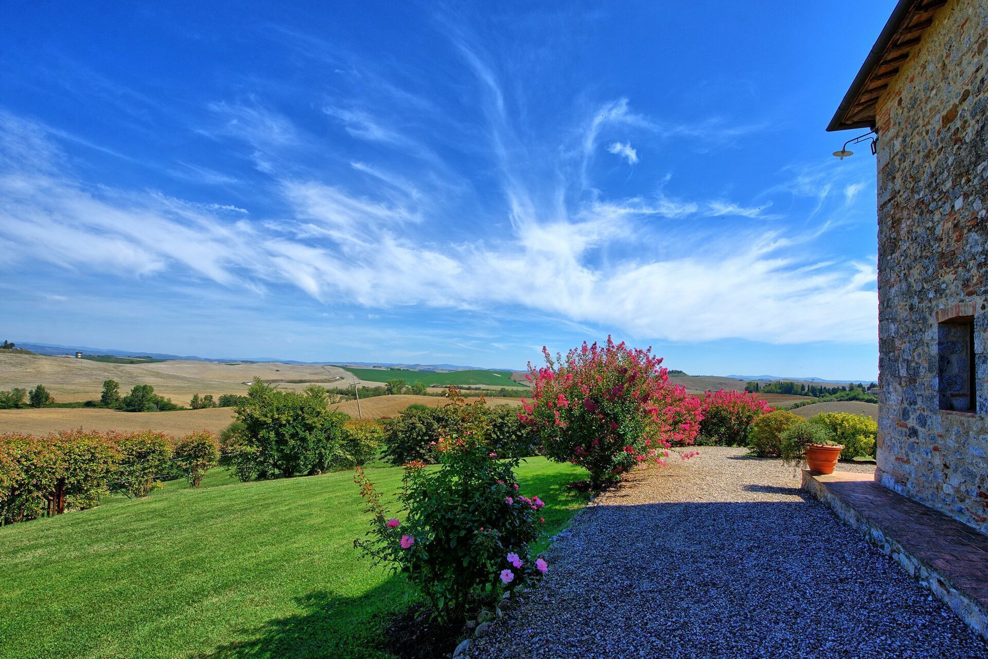 A stone building with a gravel pathway leading to a lush green lawn and flowering bushes.