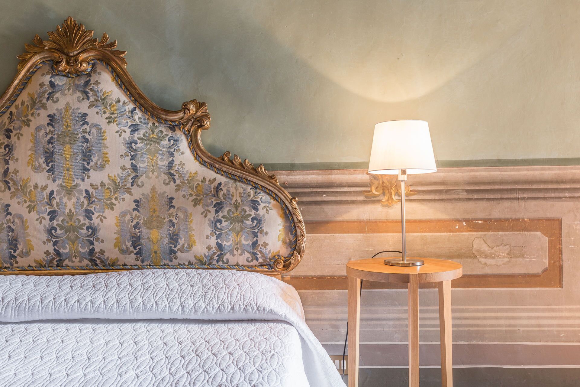 A bed with a floral headboard, a round wooden nightstand with a lamp, and a wall with a decorative molding.