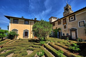 Un bâtiment historique doté d’une tour, entouré de jardins bien entretenus et agrémenté de haies aux formes géométriques.