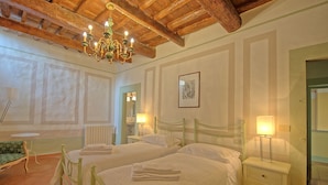Quarto de hotel com duas camas, um candelabro, uma mesa de cabeceira com abajur, um radiador e um quadro emoldurado na parede.
