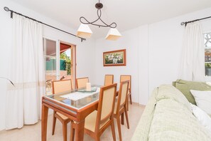 Un comedor con una mesa de vidrio y sillas de madera, una cama con cubierta verde, y una lámpara en el techo.