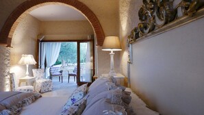 Una camera da letto con un letto grande, due sedie, un tavolo e una lampada. Un arco conduce a una zona esterna con un tavolo e sedie.