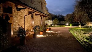Un edificio in pietra con un'area esterna coperta, piante in vaso e un vialetto sterrato che conduce a un ingresso illuminato.