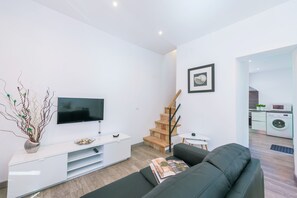 A modern living room with a flat-screen TV, a sofa, a white entertainment unit, and a staircase leading to a kitchen area.