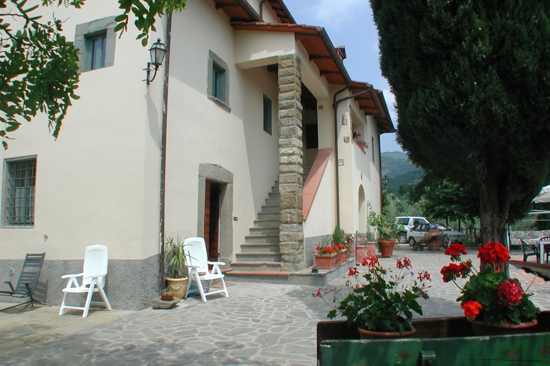 A building with a stone staircase leading to a door, white chairs outside, and potted plants with red flowers.
