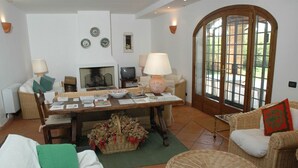 A cozy living room with a fireplace, a dining table, wicker furniture, and a large arched window.