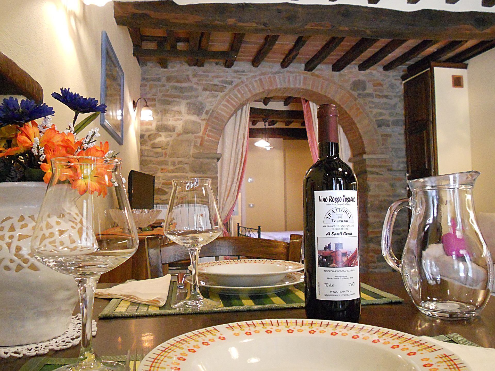 A dining table set with wine glasses, a bottle of wine, and a pitcher, with a stone wall and wooden beams in the background.