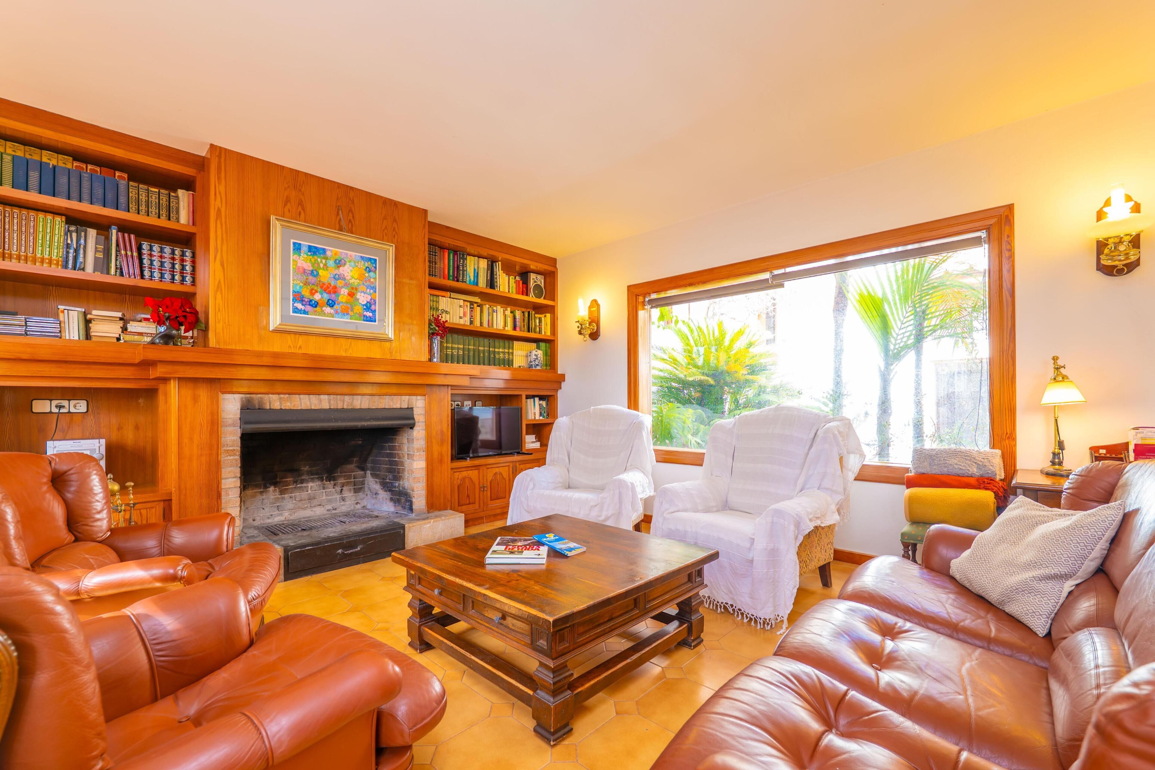Una sala de estar con sofás de cuero, una chimenea, una estantería y un ventanal con vista a palmeras.