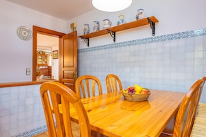 A dining area with a wooden table and chairs, a fruit basket, and a kitchen visible in the background.