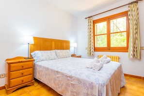 A bedroom with a wooden headboard, a single bed with floral bedding, a nightstand with a lamp, a window with curtains, and a radiator.