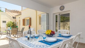 Un coin repas avec une table dressée pour quatre personnes, comprenant un saladier de fruits, des assiettes et des couverts. Une fenêtre offre une vue sur un bâtiment à l’extérieur.