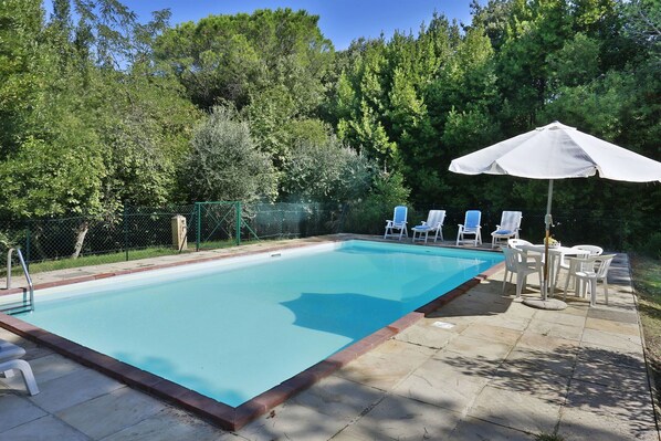 A rectangular swimming pool with a tiled edge, surrounded by a stone deck. There are white lounge chairs and a white umbrella on the deck.