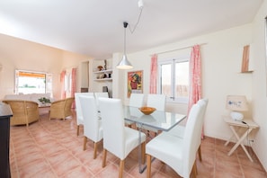 Een eethoek met een glazen tafel, witte stoelen, een hanglamp en uitzicht op buiten.
