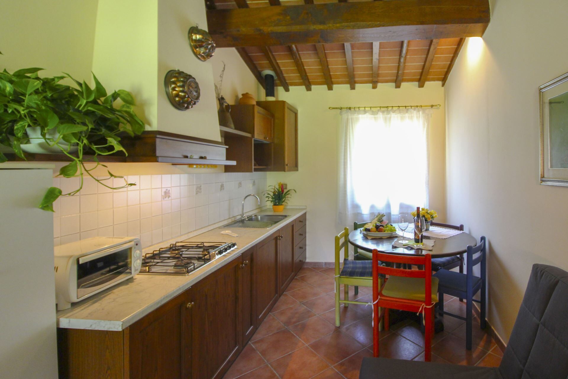 A compact kitchen with a gas stove, a sink, and wooden cabinets. There is a dining area with a table and chairs, and a window with curtains.