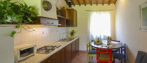 Uma cozinha compacta com fogão a gás, pia e armários de madeira. Há uma área de refeições com mesa e cadeiras, e uma janela com cortinas.