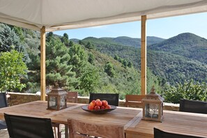 A wooden table with a bowl of fruit, two lanterns, and chairs under a canopy with a mountain view.