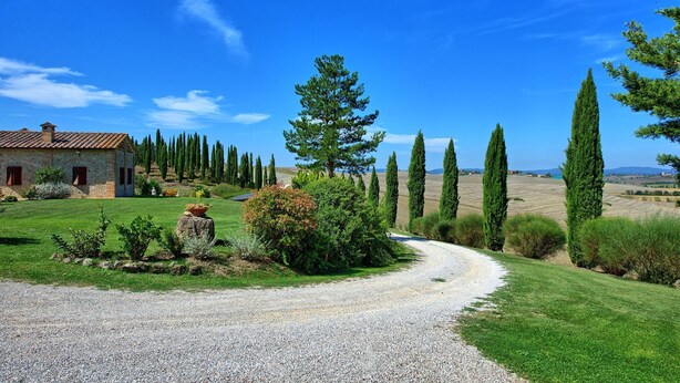 A rural landscape with a gravel path leading to a stone house with a tiled roof.