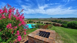 Una piscina con una griglia barbecue in mattoni, circondata da vegetazione lussureggiante e fiori rosa, con vista panoramica su dolci colline e un cielo sereno.