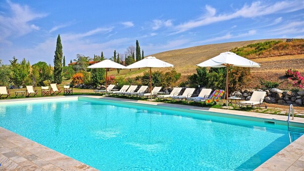 A swimming pool with lounge chairs and umbrellas, surrounded by greenery and hills.