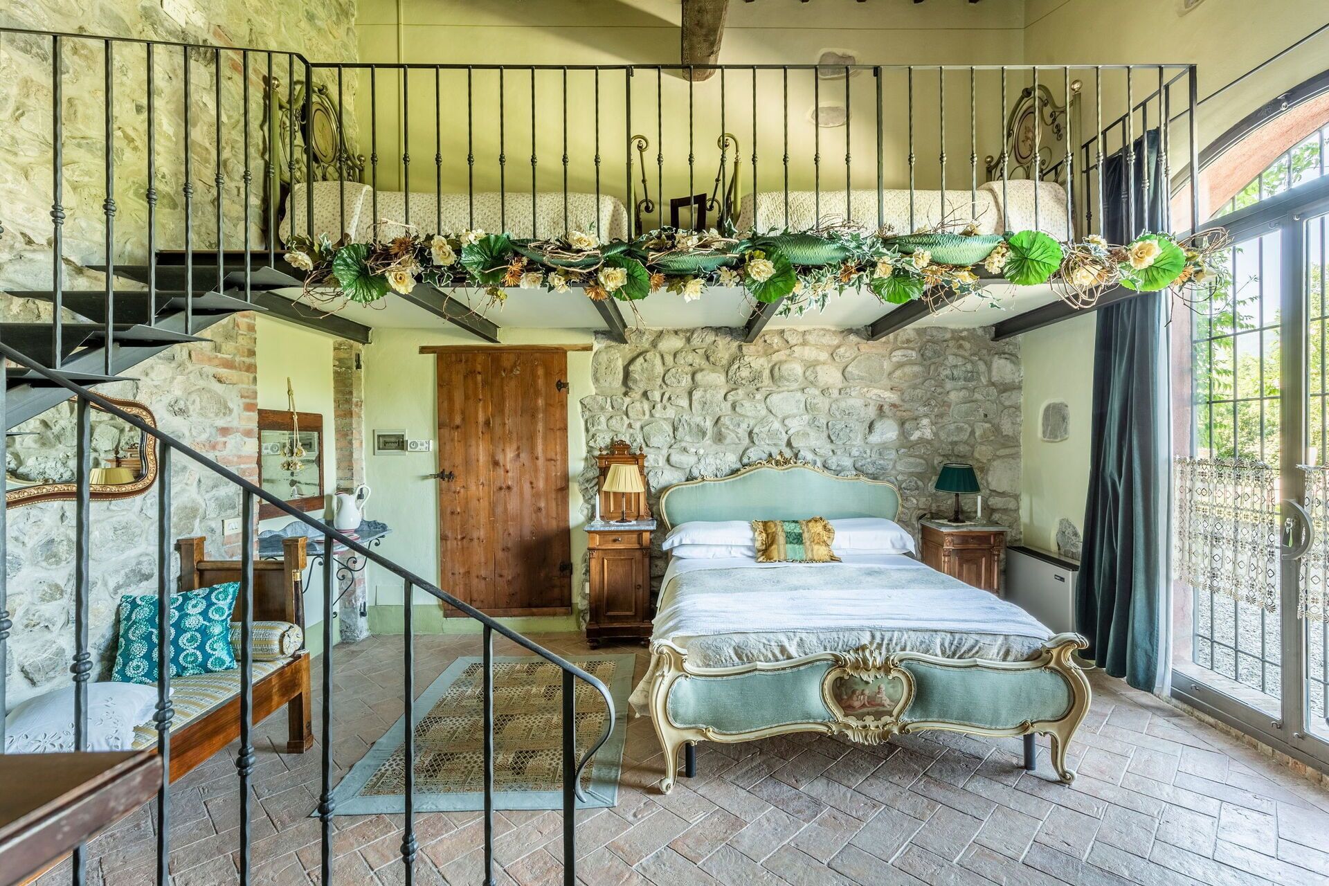 Quarto com parede de pedra, cama com cabeceira ornamentada, escada com corrimão em ferro forjado e porta de madeira.