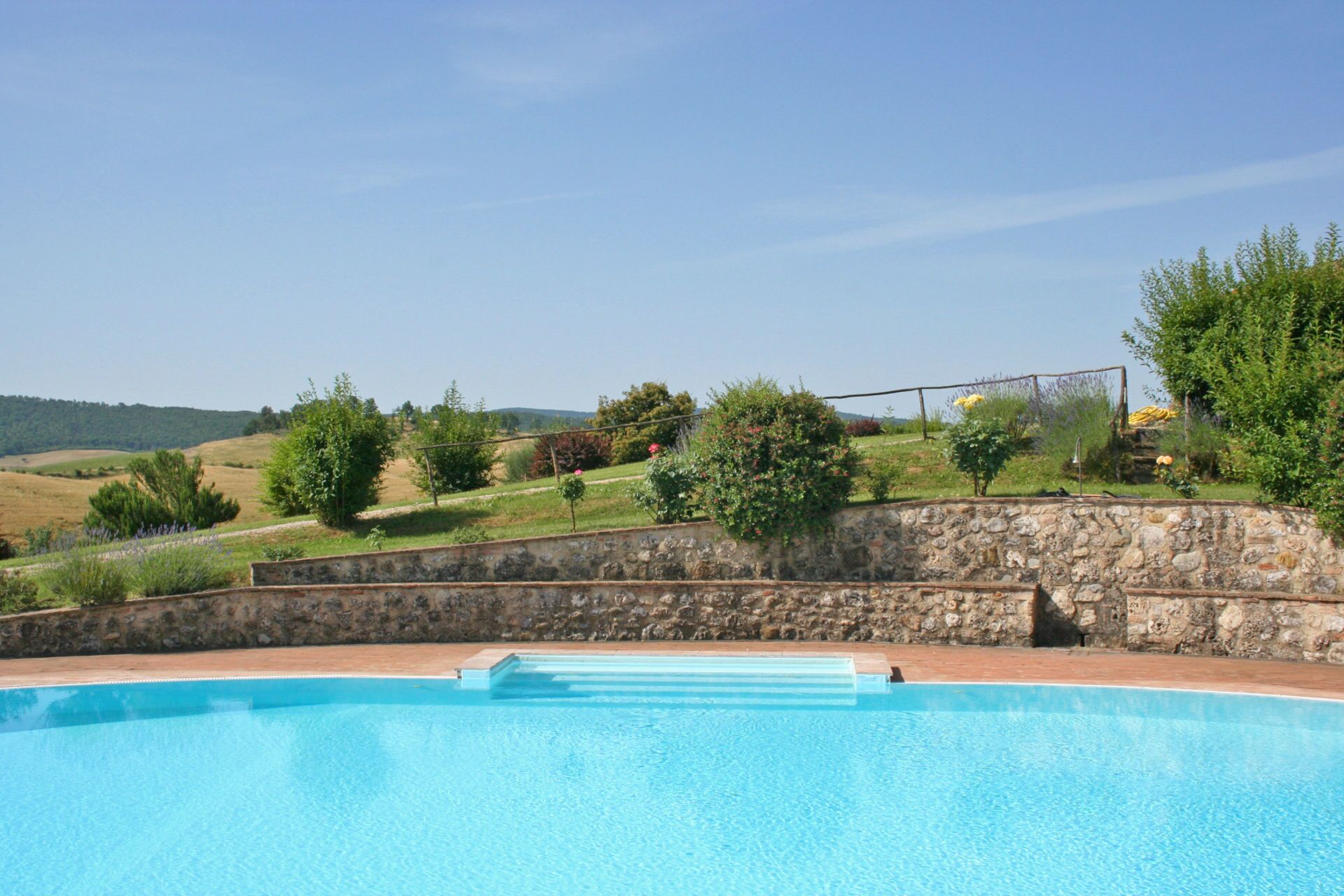 A swimming pool with a stone wall and steps leading up to it, surrounded by greenery and hills in the background.