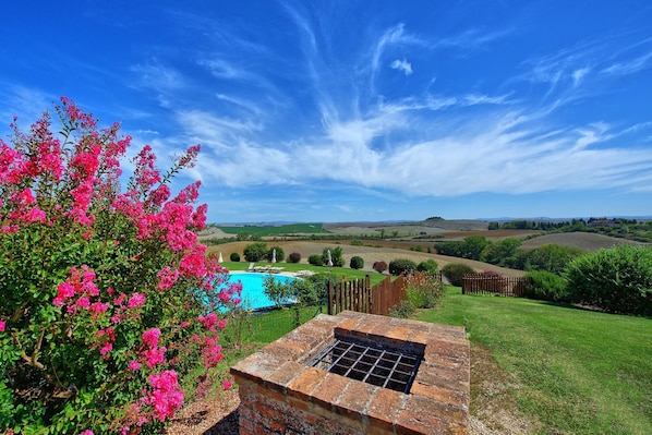 A pool with a brick barbecue grill, surrounded by lush greenery and pink flowers, with a scenic view of rolling hills and a clear sky.