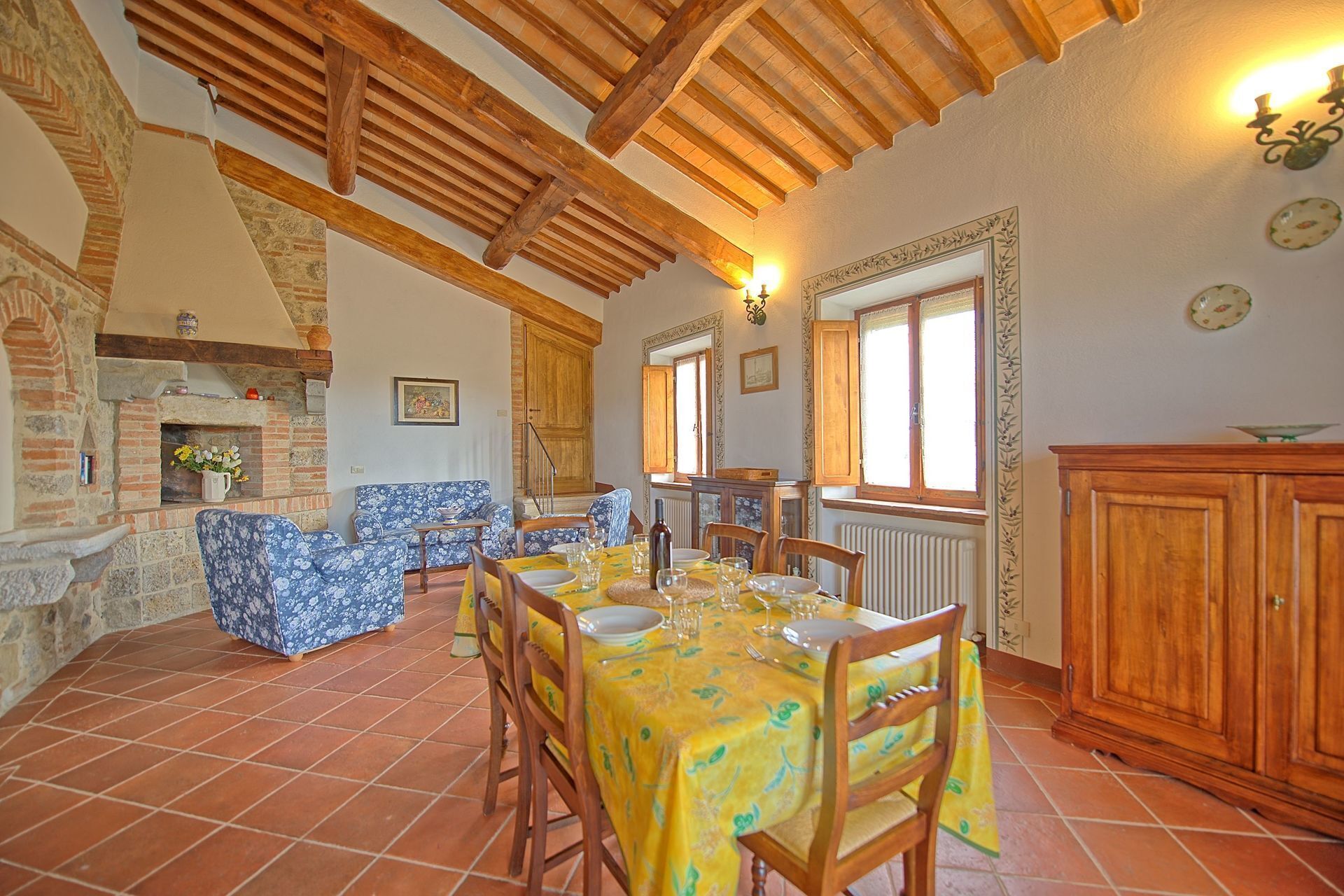 A dining area with a tiled floor, wooden ceiling beams, a fireplace, and a dining table set for six.