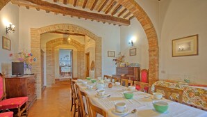 Uma sala de jantar com uma mesa comprida posta para refeição, cadeiras de madeira e uma televisão em um móvel.