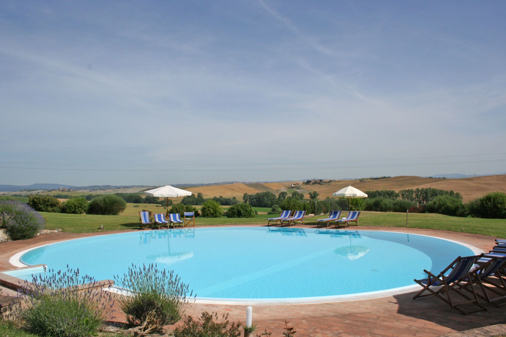 Ein großer, klarer, blauer Pool mit Liegestühlen und Sonnenschirmen, umgeben von einem angelegten Garten und Hügeln im Hintergrund.