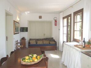 A dining area with a wooden table, chairs, a fruit basket, a sofa with patterned cushions, and a television mounted on the wall. - Nice apartment for 4 guests with WIFI, pool, TV and patio (Sassetta)