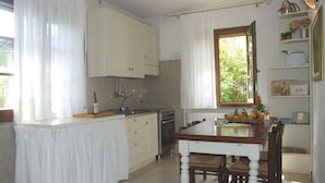 Una cocina con armarios blancos, una mesa de comedor, sillas, un ventanal con cortinas y vista a follaje exterior.