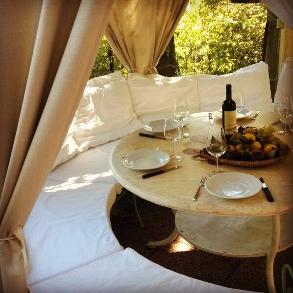 Une table ronde dressée pour un repas, avec des assiettes, des couverts, du vin et un panier de fruits.