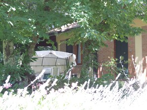A covered outdoor seating area with a table and chairs, surrounded by trees and plants.