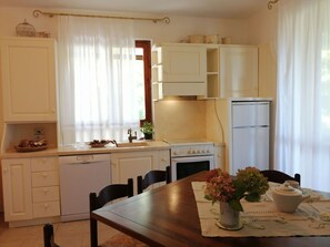 A compact kitchen with white cabinets, a stove, and a refrigerator. There is a dining table with chairs and a vase of flowers. - Beautiful apartment for 5 guests with pool, WIFI, TV and patio (Sassetta)