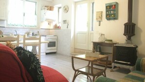A cozy living room with a red sofa, a green armchair, and a wooden table. There is a kitchen area with white cabinets and a stove. A clock and a shelf with decorative items are also visible. - Nice apartment for 6 guests with WIFI and TV (Sassetta)
