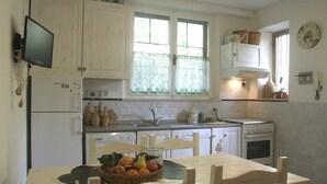 A kitchen with white cabinets, a wooden dining table with chairs, a fruit basket, and a window with lace curtains. - Nice apartment for 6 guests with WIFI and TV (Sassetta)