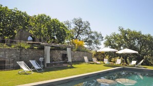 Piscina con sillas de descanso y sombrillas, rodeada por un muro de piedra y árboles.