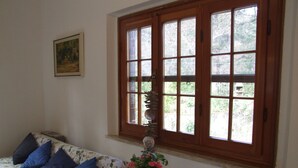 Una habitación con ventana de madera, un sofá floral con cojines azules, una mesita redonda con mantel floral y una planta en maceta con flores rosas.